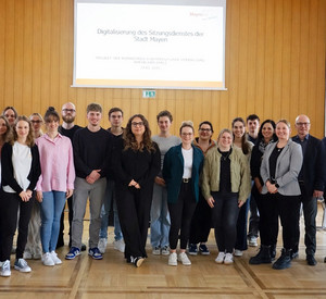 Gruppenfoto in einem großen Raum. Etwa 20 Personen, darunter Lehrgangsteilnehmende der ZVS RLP und Mitarbeitende der Stadtverwaltung Mayen, stehen nebeneinander und schauen in die Kamera. Im Hintergrund ist eine Leinwand mit der Präsentationsfolie „Digitalisierung des Sitzungsdienstes der Stadt Mayen“ zu sehen.
